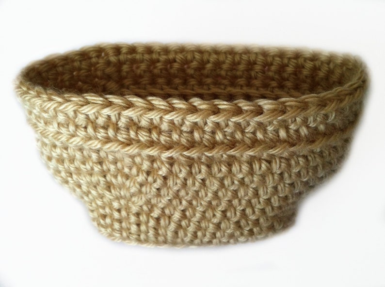 May include: A hand-crocheted basket in a light tan color. The basket has a rounded shape with a wide opening and a textured surface. The stitches are clearly visible, creating a detailed pattern. This item is suitable for home decor.