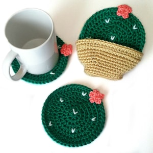May include: Handmade crochet cactus coasters in green with white accents and small pink flowers. One coaster is under a white mug, and another is paired with a tan crochet pot. The coasters are round and add a decorative touch to any home.