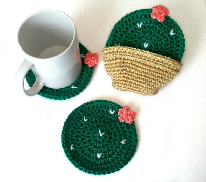 May include: Handmade crochet cactus coasters in green with white accents and small pink flowers. One coaster is under a white mug, and another is paired with a tan crochet pot. The coasters are round and add a decorative touch to any home.