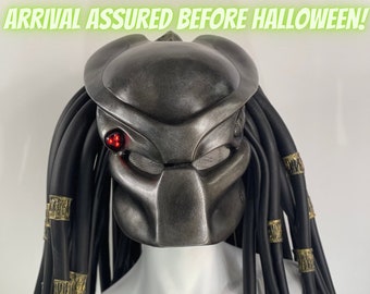 May include: A Predator mask with a metallic silver finish, featuring a red targeting device and black dreadlocks adorned with gold accents. The text "ARRIVAL ASSURED BEFORE HALLOWEEN!" is displayed above the mask.