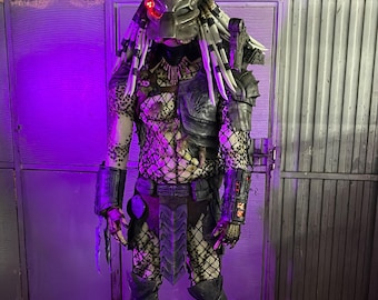 May include: A full-body Predator costume with a detailed mask, dreadlocks, and armor. The costume features a mesh body suit, intricate detailing, and clawed hands and feet. The Predator's mask has a red targeting system.
