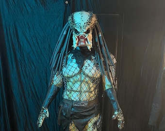 May include: A Predator costume with a black and green patterned body suit, a black leather belt, and a detailed mask with dreadlocks.
