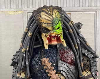 May include: A Predator action figure with a black and silver armored suit, a bone-like weapon, and a green and yellow mask with a menacing expression.