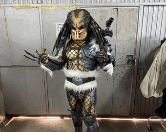 May include: A Predator costume with a black and gray patterned design. The costume includes a mask, a chest piece, a belt, and leg coverings. The Predator is holding a weapon in its right hand.