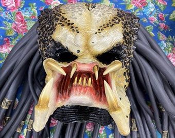 May include: A black and yellow Predator mask with dreadlocks. The mask has a red mouth with sharp teeth and gold accents.
