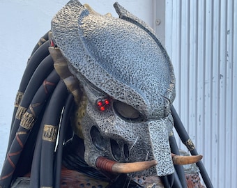 Pode incluir: Uma máscara de Predator com um design texturizado cinza e marrom. A máscara tem olhos vermelhos brilhantes e duas presas brancas longas.