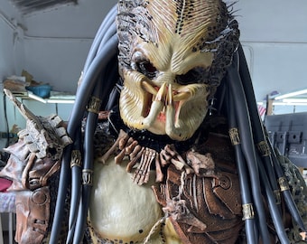 Pode incluir: Um traje detalhado do Predador com um design texturizado, semelhante a um réptil, em tons de bege, castanho e cinza. A máscara tem mandíbulas proeminentes e dreadlocks. A armadura do peito e dos ombros é intrincadamente detalhada, com detalhes em osso e couro.