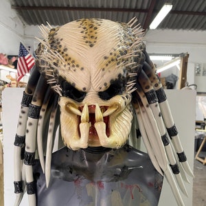 May include: A detailed Predator mask with a white and brown camouflage pattern, sharp teeth, and dreadlocks. The mask is on a mannequin torso.