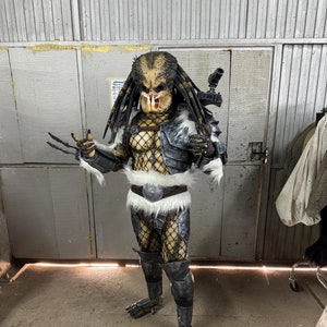 May include: A Predator costume with a black and gray patterned design. The costume includes a mask, a chest piece, a belt, and leg coverings. The Predator is holding a weapon in its right hand.