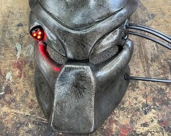 May include: A silver Predator mask with a red light on the side. The mask has a detailed design and a textured surface.