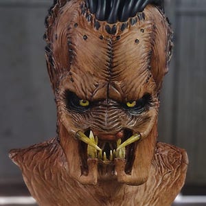 Puede incluir: Una detallada máscara de Depredador marrón con ojos amarillos y dientes afilados. La máscara tiene una superficie texturizada y está montada sobre una base negra, mostrando los intrincados detalles del diseño alienígena.