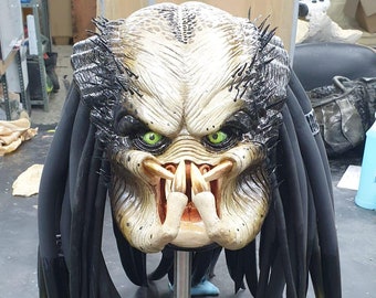 Pode incluir: Um close-up de uma máscara de Predator com dreadlocks pretos. A máscara é feita de um material marrom claro e tem olhos verdes. A máscara está em um suporte preto.