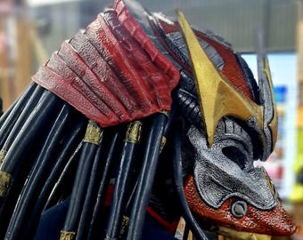 May include: A red and gold Predator mask with black dreadlocks. The mask has a detailed design with sharp teeth and a textured surface.