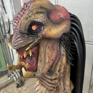 Könnte beinhalten: Eine Nahaufnahme einer Predator-Maske mit einem roten Auge, scharfen Zähnen und schwarzen Dreadlocks. Die Maske ist aus einem strukturierten Material gefertigt und wirkt realistisch.