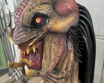 May include: A close-up of a Predator mask with a red eye, sharp teeth, and black dreadlocks. The mask is made of a textured material and has a realistic appearance.