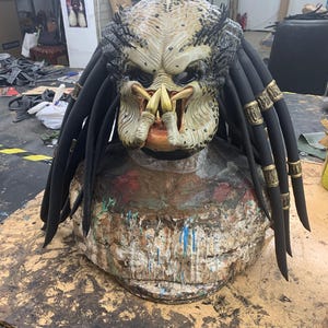 Könnte beinhalten: Eine Predator-Maske mit Dreadlocks, aus hellem braunen Material mit schwarzen und gelben Details. Die Maske hat einen bedrohlichen Ausdruck mit scharfen Zähnen und einem einzigen roten Auge.