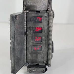 May include: A detailed, dark gray prop with a hinged door and multiple red illuminated displays. The prop has a textured surface and appears to be a futuristic device or gadget. The displays show red symbols, adding to the sci-fi aesthetic.