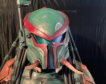 May include: A green and red Predator helmet with dreadlocks and tusks. The helmet has a clear visor and a green and red design.