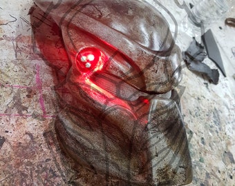 Pode incluir: Uma máscara de Predator marrom e cinza com olhos vermelhos brilhantes. A máscara é feita de um material plástico duro e tem um design detalhado.