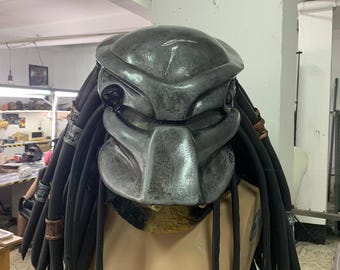 May include: A gray and black Predator mask with dreadlocks. The mask has a detailed design with a textured surface and a mesh visor.