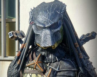 Pode incluir: Um traje de Predator com uma máscara preta e prateada, dreadlocks pretos e uma peça de peito preta e prateada com uma correia de couro marrom.