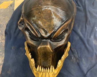 Könnte beinhalten: Eine detaillierte Predator-Maske in Bronze mit einem goldfarbenen Unterkiefer und scharfen Zähnen. Die Maske ist aus einem harten, strapazierfähigen Material gefertigt und hat ein realistisches Aussehen.