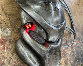 Puede incluir: Una máscara de Predator plateada con luces rojas brillantes. La máscara tiene un aspecto desgastado y deteriorado, con arañazos y marcas visibles. La máscara está hecha de un material plástico duro y tiene un diseño detallado.