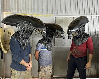 May include: Three people wearing black alien-like masks with detailed textured surfaces. The masks are worn over their heads and cover their faces. The people are wearing casual clothing.