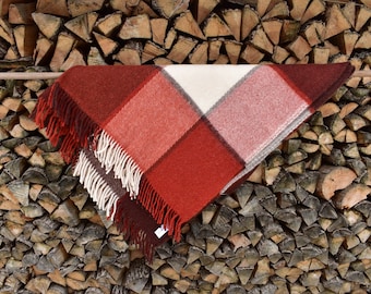 May include: A red, white, and brown plaid throw blanket with fringe on the edges. The blanket is hanging on a wooden rod in front of a wall of stacked firewood.