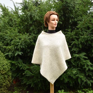 May include: A white knitted poncho with a simple design. The poncho is draped over a mannequin and is shown against a backdrop of green foliage.