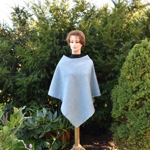 Könnte beinhalten: Ein hellblauer Strickponcho mit Rippenstruktur. Der Poncho ist über eine Schaufensterpuppe gelegt und vor einem Hintergrund aus grünem Laub dargestellt.