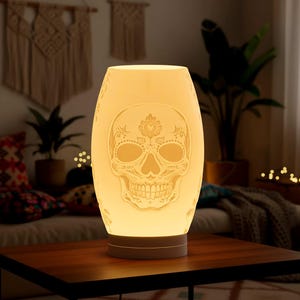 Puede incluir: Lámpara en forma de calavera de color crema brillante, con intrincados detalles florales y de calaveras. La lámpara tiene forma ovalada y se apoya sobre una base redonda gris. Está sobre una mesa de madera en una habitación tenue.