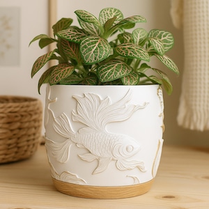 May include: A white ceramic planter with a detailed relief of a goldfish. The planter has a light wood-colored base and is filled with a green plant with pink-veined leaves. The fish design is a prominent feature of the pot.