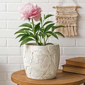 May include: A white ceramic flower pot with a detailed floral relief design, holding a peony plant with a large pink flower and green leaves. The pot sits on a round wooden table, creating a decorative display.