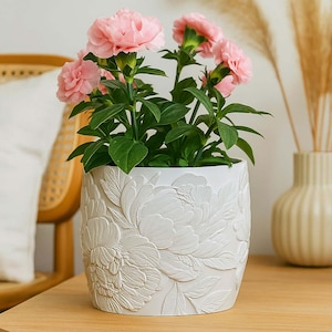 Puede incluir: Una maceta de cerámica blanca con un diseño floral en relieve detallado, llena de claveles rosas vibrantes y follaje verde. La maceta se encuentra sobre una superficie de madera, mostrando el contraste entre la maceta texturizada y las flores lisas.