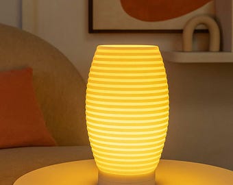 Puede incluir: Lámpara de mesa amarilla con pantalla cilíndrica y acanalada. La lámpara emite una luz cálida, creando un ambiente acogedor. Se encuentra sobre una mesa blanca, con un fondo borroso.