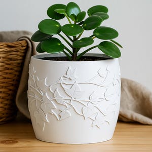 Puede incluir: Una maceta de cerámica blanca con un diseño de hojas de hiedra en relieve, que contiene una planta verde con hojas redondas. La maceta está sobre una superficie de madera, con una cesta de mimbre y tela beige en el fondo.