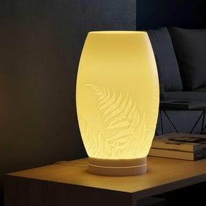 May include: A glowing, off-white ceramic table lamp with a fern leaf design. The lamp has a cylindrical shape with a wider middle and a smaller base. The fern pattern is etched into the surface, and the lamp emits a warm, yellow light.
