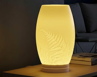 May include: A glowing, off-white ceramic table lamp with a fern leaf design. The lamp has a cylindrical shape with a wider middle and a smaller base. The fern pattern is etched into the surface, and the lamp emits a warm, yellow light.