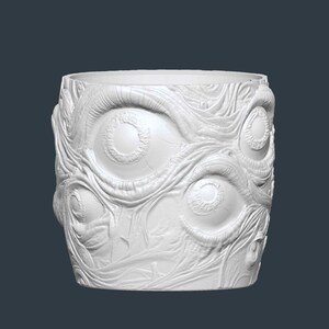 May include: White cylindrical plant pot with a textured surface featuring multiple eye designs. The pot has a wide opening and is made of a solid, opaque material. The design is a unique, artistic representation.