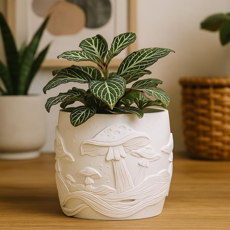 May include: A white ceramic planter with a raised mushroom design, holding a green plant with white veined leaves. The planter has a cylindrical shape and is sitting on a wooden surface. The background includes a framed artwork and a woven basket.