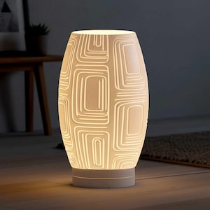 May include: A white ceramic table lamp with a geometric pattern of recessed squares and rectangles. The lamp has a cylindrical shape with a wider base and a glowing interior. The lamp is on a white base.