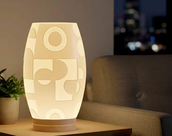 Puede incluir: Una lámpara de mesa color crema con un patrón geométrico de círculos y cuadrados. La lámpara tiene forma cilíndrica y se apoya sobre una base marrón claro. La lámpara está encendida, emitiendo un brillo suave, y está colocada sobre una mesa de madera.