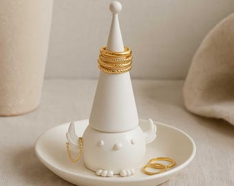 May include: A white ceramic bird-shaped ring holder with a conical top and a small dish base. Several gold rings are stacked on the top. Additional gold rings rest in the dish. A gold chain hangs from the bird's wing.