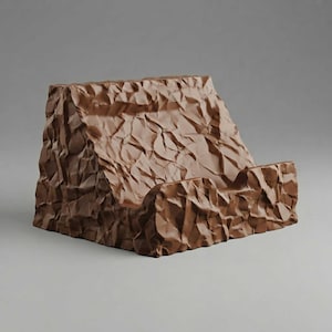 May include: A brown phone stand featuring a textured, crinkled surface that mimics the appearance of crumpled paper. The stand has a slanted backrest and a small front ledge designed to hold a mobile device in a stable, upright position.