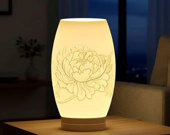 May include: A cream-colored ceramic table lamp with a floral design. The lamp has an oval shape and a carved peony flower on the front. The lamp is lit, casting a warm glow. It sits on a wooden table.
