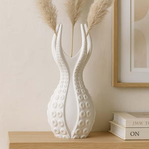 May include: A white ceramic vase with a unique design, featuring two curved arms that extend upwards. The vase has a textured surface with circular patterns and holds dried pampas grass. The vase is on a wooden surface.