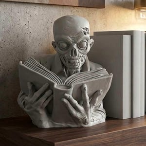 May include: A grey zombie bookend, depicting a zombie reading a book. The sculpture has detailed features and is placed on a wooden surface. A stack of books is visible in the background, adding to the decorative element.
