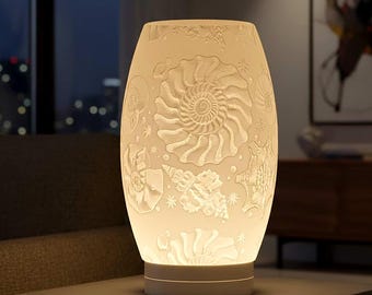 May include: A white, oval-shaped lamp with a detailed relief pattern of seashells and aquatic designs. The lamp is illuminated from within, casting a warm glow. It sits on a white base, creating a soft, ambient light.