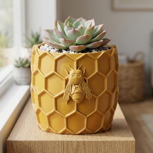Può includere: Un vaso cilindrico color giallo senape caratterizzato da un motivo a nido d'ape in rilievo sulla superficie esterna. Una dettagliata figura di ape tridimensionale è centrata sulla parte anteriore. Il vaso contiene una pianta succulenta.
